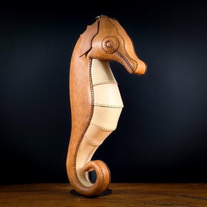 May include: A handcrafted leather seahorse sculpture in shades of tan and light beige. The seahorse is meticulously stitched, with a detailed head and a curled tail. It stands upright on a small, round base, showcasing the craftsmanship of the leatherwork.