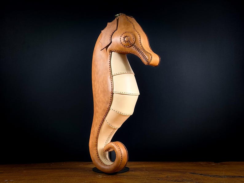 May include: A handcrafted leather seahorse sculpture in shades of tan and light beige. The seahorse is meticulously stitched, with a detailed head and a curled tail. It stands upright on a small, round base, showcasing the craftsmanship of the leatherwork.