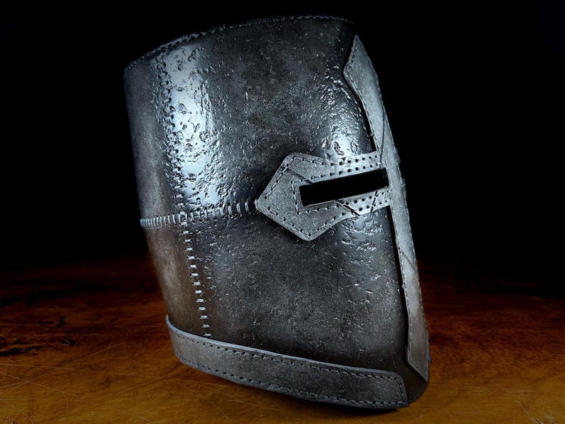K&ouml;nnte beinhalten: Ein verwittert aussehender Helm in Silber-Grau mit Kreuzdesign und horizontalem Augenschlitz. Der Helm weist sichtbare N&auml;hte und eine strukturierte Oberfl&auml;che auf, was auf ein mittelalterliches oder historisches Thema hindeutet.