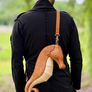 May include: A unique, handcrafted leather crossbody bag shaped like a seahorse. The bag features a tan and cream color scheme with a long, adjustable strap. The seahorse design includes detailed stitching and metal hardware.