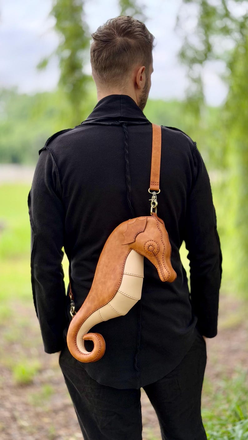 May include: A unique, handcrafted leather crossbody bag shaped like a seahorse. The bag features a tan and cream color scheme with a long, adjustable strap. The seahorse design includes detailed stitching and metal hardware.
