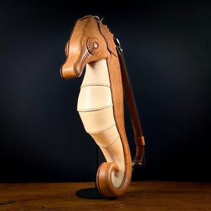 May include: A handcrafted leather seahorse-shaped bag in shades of tan and brown. The bag features a long, adjustable strap and detailed stitching. The seahorse design includes a head, body, and tail, with a unique, artistic flair.