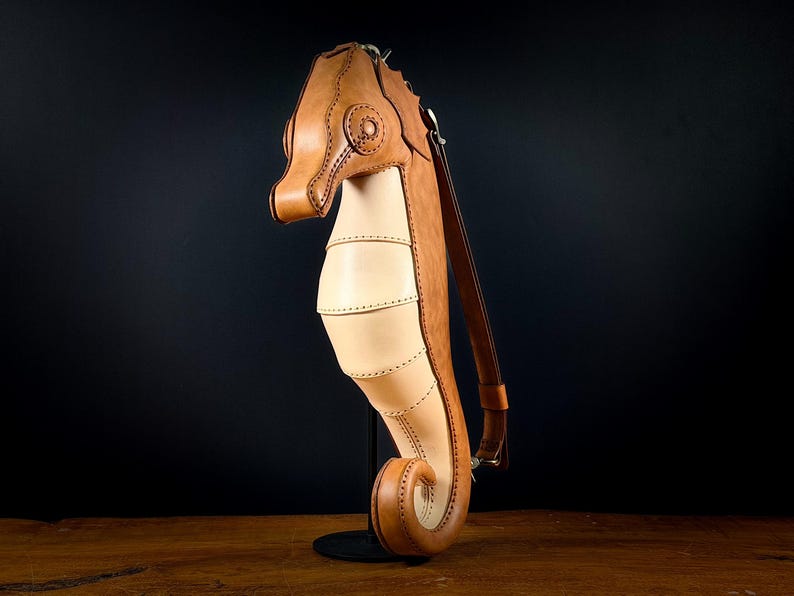 May include: A handcrafted leather seahorse-shaped bag in shades of tan and brown. The bag features a long, adjustable strap and detailed stitching. The seahorse design includes a head, body, and tail, with a unique, artistic flair.