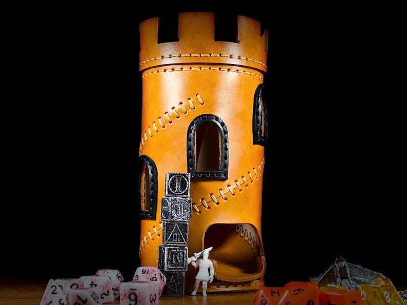 May include: A tan leather dice tower, crafted to resemble a castle turret. It has stitched seams, arched windows, and a dice-rolling opening. Dice and a miniature figure are placed near the tower.