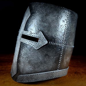 K&ouml;nnte beinhalten: Ein silberfarbener mittelalterlicher Helm mit Kreuzdesign und rechteckigem Augenschlitz. Der Helm hat eine strukturierte Oberfl&auml;che und sichtbare N&auml;hte. Der dunkle Hintergrund hebt die Details des Helms hervor.