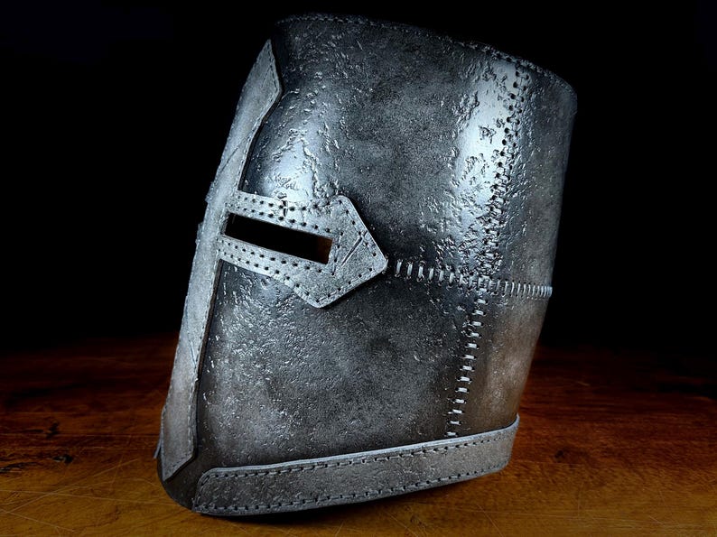 K&ouml;nnte beinhalten: Ein silberfarbener mittelalterlicher Helm mit Kreuzdesign und rechteckigem Augenschlitz. Der Helm hat eine strukturierte Oberfl&auml;che und sichtbare N&auml;hte. Der dunkle Hintergrund hebt die Details des Helms hervor.