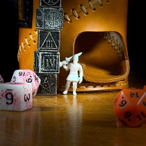 May include: A close-up of a tabletop scene with a brown leather dice tower, a stack of black dice, and various polyhedral dice. A small white figurine with a sword stands near the tower. Some dice are white with red splatters.