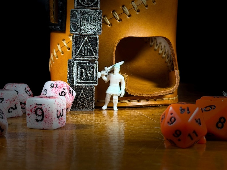 May include: A close-up of a tabletop scene with a brown leather dice tower, a stack of black dice, and various polyhedral dice. A small white figurine with a sword stands near the tower. Some dice are white with red splatters.
