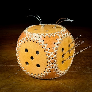 May include: A tan leather pincushion shaped like a die, with six sides. The edges are stitched with white thread. Several needles are inserted into the sides. The pincushion has black dots on each side, representing the numbers on a die.