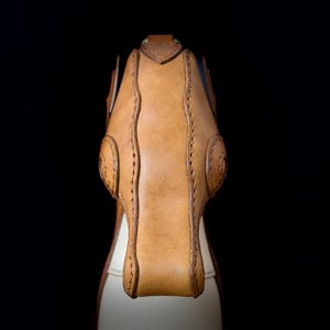 May include: Close-up of a handcrafted leather bag, showcasing a tan exterior with intricate stitching and a contrasting off-white interior. The bag features a structured design with rounded details and a secure closure, set against a black backdrop.