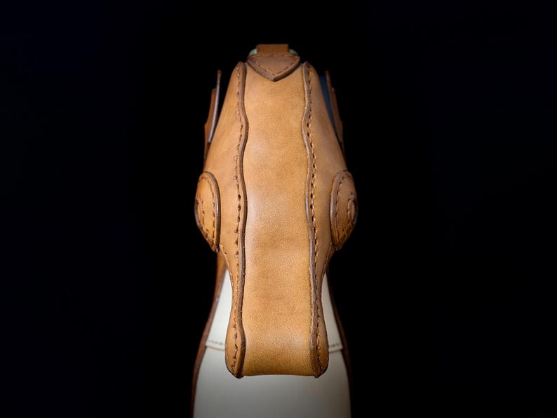 May include: Close-up of a handcrafted leather bag, showcasing a tan exterior with intricate stitching and a contrasting off-white interior. The bag features a structured design with rounded details and a secure closure, set against a black backdrop.