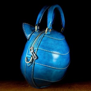May include: A handcrafted, blue leather handbag with a unique, rounded shape and a top handle. The bag features white stitching, a zipper closure, and a decorative charm. The bag is set against a dark background.