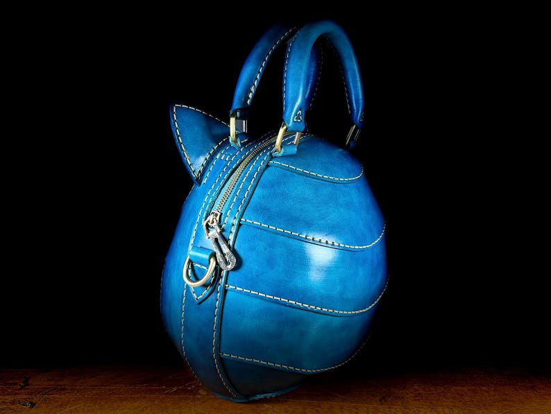 May include: A handcrafted, blue leather handbag with a unique, rounded shape and a top handle. The bag features white stitching, a zipper closure, and a decorative charm. The bag is set against a dark background.