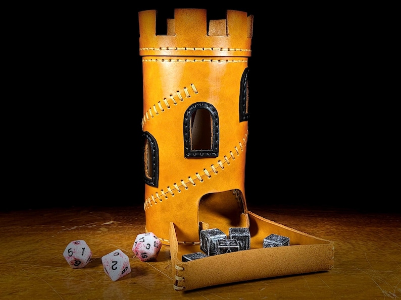 May include: A tan leather dice tower, resembling a castle, with black window frames and decorative stitching. The tower has a tray for dice to roll into, and several dice are scattered around it.
