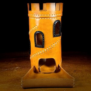May include: A tan leather dice tower, resembling a castle, with black window details and contrasting stitching. The tower has a matching tray at the base. The background is dark. The tower is designed for tabletop gaming.
