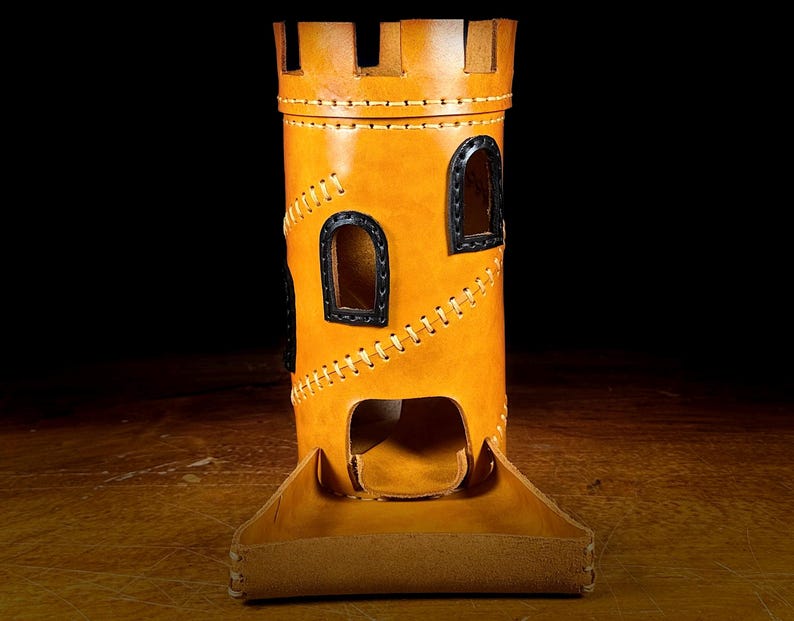 May include: A tan leather dice tower, resembling a castle, with black window details and contrasting stitching. The tower has a matching tray at the base. The background is dark. The tower is designed for tabletop gaming.