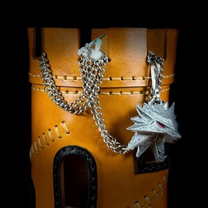 May include: A tan leather container with a silver chain and a silver wolf head pendant. The container has black trim and decorative stitching. The background is black.
