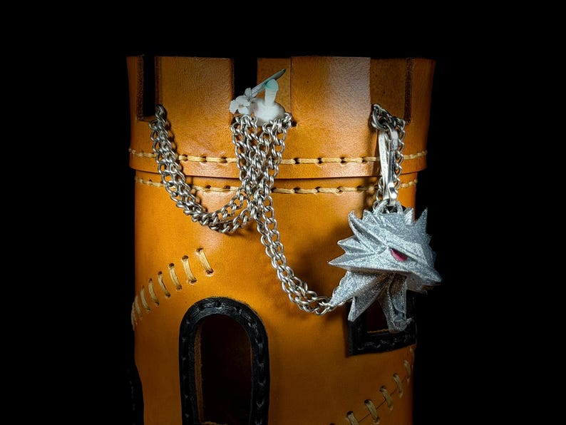 May include: A tan leather container with a silver chain and a silver wolf head pendant. The container has black trim and decorative stitching. The background is black.