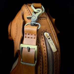 May include: Close-up of a brown leather bag with a brass zipper and buckle. The bag has a shoulder strap with a metal clasp. The leather is stitched with a contrasting thread, and the bag has a textured surface.