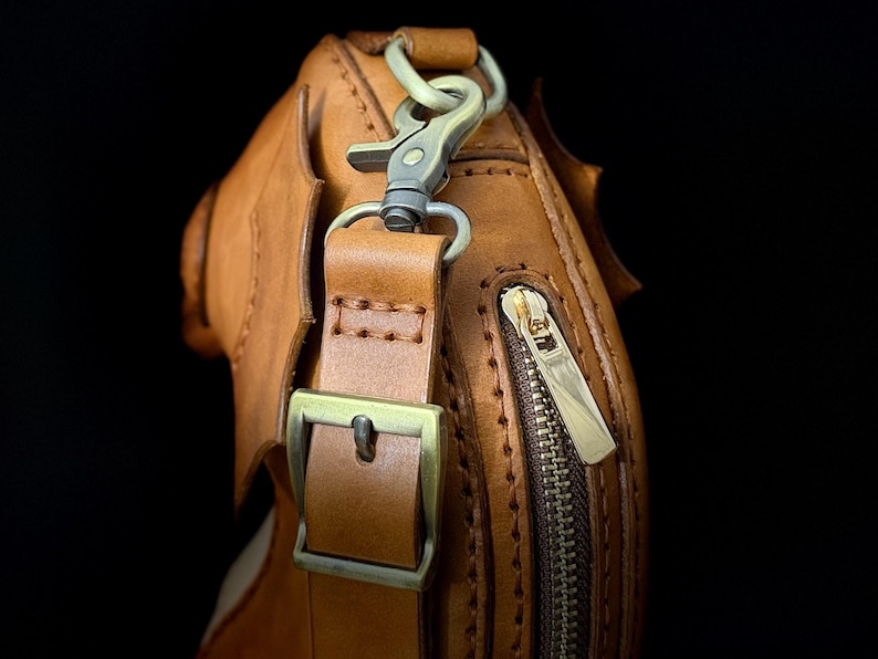 May include: Close-up of a brown leather bag with a brass zipper and buckle. The bag has a shoulder strap with a metal clasp. The leather is stitched with a contrasting thread, and the bag has a textured surface.
