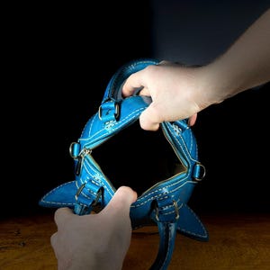 May include: A bright blue leather handbag with white stitching and silver-tone hardware. The bag has a zipper closure and two handles. The bag is held open, revealing the interior. The background is dark.