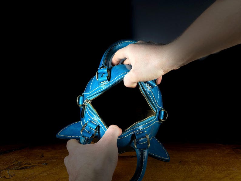 May include: A bright blue leather handbag with white stitching and silver-tone hardware. The bag has a zipper closure and two handles. The bag is held open, revealing the interior. The background is dark.