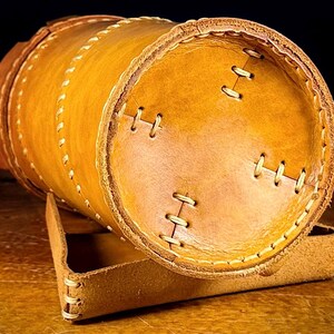 May include: A cylindrical, handmade leather container in shades of tan and brown, with visible stitching and a matching leather stand. The container is approximately 15 cm long and 10 cm in diameter, showcasing a rustic aesthetic.