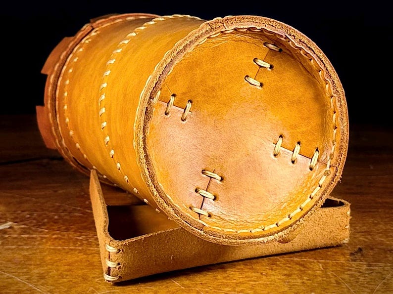 May include: A cylindrical, handmade leather container in shades of tan and brown, with visible stitching and a matching leather stand. The container is approximately 15 cm long and 10 cm in diameter, showcasing a rustic aesthetic.