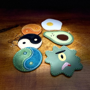 Puede incluir: Colección de parches de cuero coloridos de varias formas. Incluye un símbolo de yin-yang en naranja y negro, un huevo frito, un aguacate y un germen verde con una cara. Cada parche tiene un borde cosido.