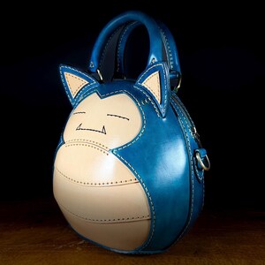 May include: A handcrafted blue and tan leather handbag shaped like a cartoon character. The bag features a top handle, a zipper closure, and a detachable shoulder strap. The design includes stitched details and a character face.