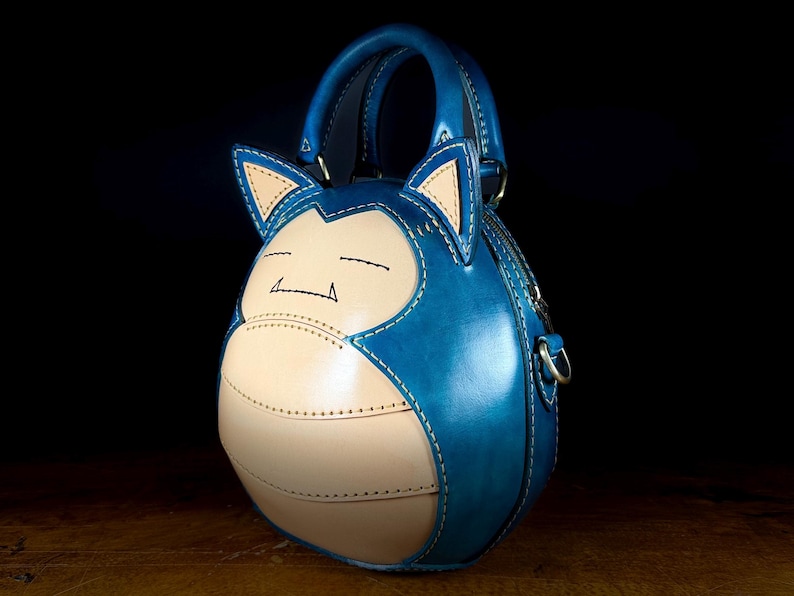May include: A handcrafted blue and tan leather handbag shaped like a cartoon character. The bag features a top handle, a zipper closure, and a detachable shoulder strap. The design includes stitched details and a character face.