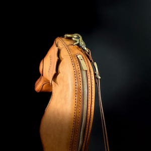 May include: A tan leather handbag with a decorative scalloped edge. The bag features a long, vertical zipper with a brass pull and a brass clasp. A thin, brown strap extends from the top of the bag.