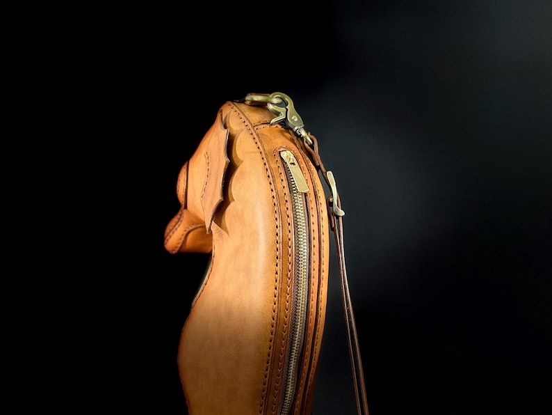 May include: A tan leather handbag with a decorative scalloped edge. The bag features a long, vertical zipper with a brass pull and a brass clasp. A thin, brown strap extends from the top of the bag.