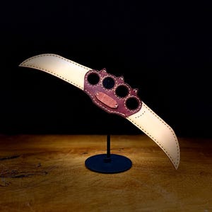 Puede incluir: Una máscara de cuero artesanal con un diseño curvo en forma de pico. La máscara presenta una sección central con tres agujeros circulares para los ojos y un emblema decorativo. El cuero es una combinación de beige y burdeos, con un soporte negro.