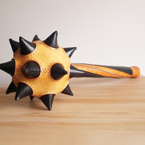 Op de afbeelding: Een lederen, met spikes bedekt kogelwapen met een zwart en bruin kleurenschema. De kogel is bedekt met zwarte spikes en heeft een bruin leren handvat.
