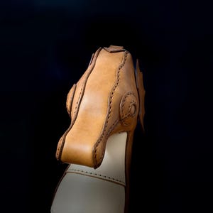 May include: Close-up of a handcrafted leather object, possibly a shoe or boot. The item features a light brown, textured leather upper with decorative stitching and a lighter-colored base. The background is a solid black.
