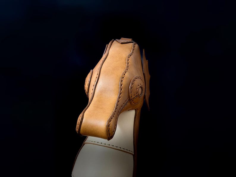 May include: Close-up of a handcrafted leather object, possibly a shoe or boot. The item features a light brown, textured leather upper with decorative stitching and a lighter-colored base. The background is a solid black.