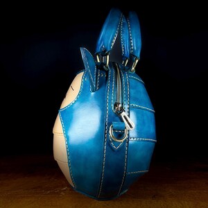 May include: A handcrafted, blue and beige leather handbag with a unique, rounded shape. The bag features a zipper closure, top handles, and white stitching details. The design incorporates a cartoon-like aesthetic, with a distinct character face.