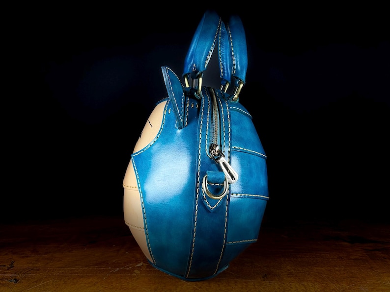 May include: A handcrafted, blue and beige leather handbag with a unique, rounded shape. The bag features a zipper closure, top handles, and white stitching details. The design incorporates a cartoon-like aesthetic, with a distinct character face.