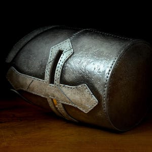 K&ouml;nnte beinhalten: Ein zylindrischer, metallisch silberner Beh&auml;lter mit strukturierter Oberfl&auml;che. Der Beh&auml;lter hat einen dekorativen, helleren silbernen Riemen mit kreuzartigem Design. Das Objekt ist vor einem dunklen Hintergrund platziert, um seine Details hervorzuheben.