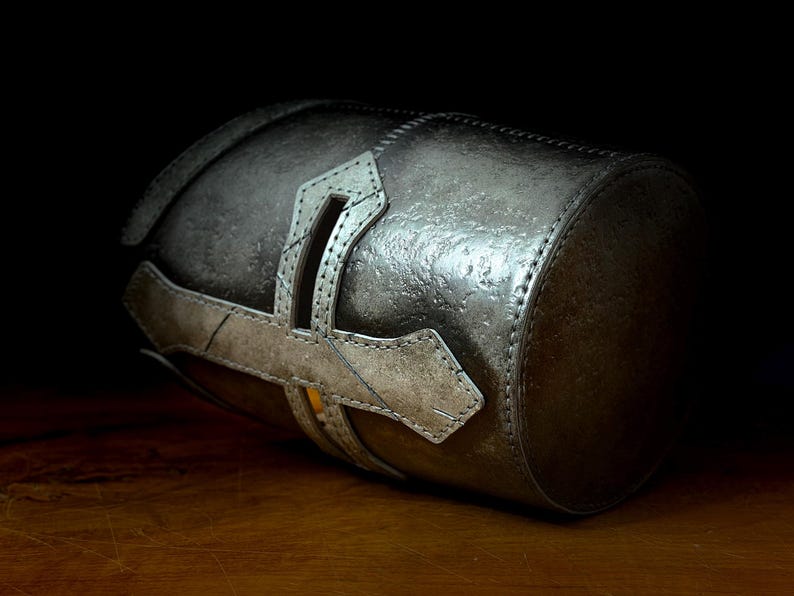 K&ouml;nnte beinhalten: Ein zylindrischer, metallisch silberner Beh&auml;lter mit strukturierter Oberfl&auml;che. Der Beh&auml;lter hat einen dekorativen, helleren silbernen Riemen mit kreuzartigem Design. Das Objekt ist vor einem dunklen Hintergrund platziert, um seine Details hervorzuheben.