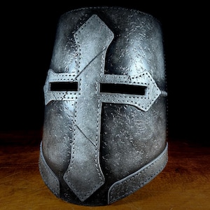 May include: A metallic gray Crusader helmet with a prominent cross design. The helmet features eye slits and is constructed with riveted panels, showcasing a medieval aesthetic. The cross is centered on the front, adding a historical and symbolic element.