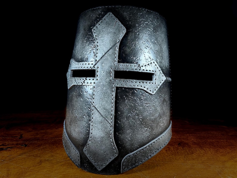 K&ouml;nnte beinhalten: Ein metallisch-grauer Kreuzritterhelm mit einem markanten Kreuzdesign. Der Helm hat Sehschlitze und ist aus genieteten Paneelen gefertigt, die eine mittelalterliche &Auml;sthetik zeigen. Das Kreuz ist mittig auf der Vorderseite angebracht.