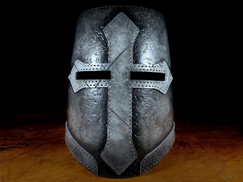 K&ouml;nnte beinhalten: Ein metallisch grauer Helm mit einem markanten Kreuzdesign. Der Helm hat eine genietete Kreuzform auf der Vorderseite, mit Augenschlitzen und einer strukturierten Oberfl&auml;che. Das Design deutet auf ein mittelalterliches oder historisches Thema hin.