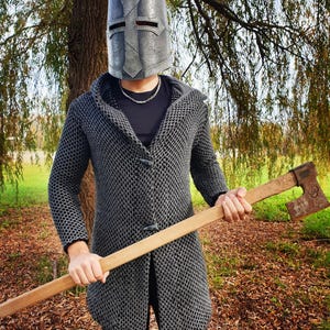 Finished leather Crusader Great Helm made from this DIY pattern. The helmet has a dark, weathered metallic finish designed to look like aged iron, worn by a person in chainmail armor holding a battle axe in a forest.