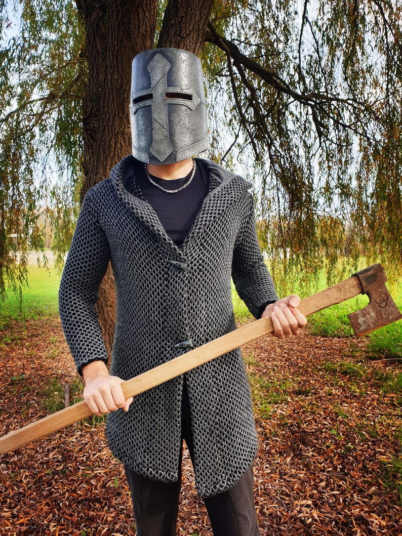 Finished leather Crusader Great Helm made from this DIY pattern. The helmet has a dark, weathered metallic finish designed to look like aged iron, worn by a person in chainmail armor holding a battle axe in a forest.