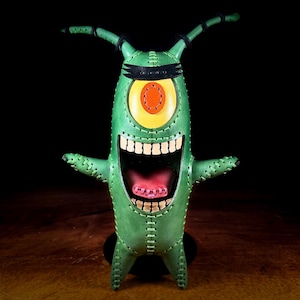 以下が含まれることがあります： 緑色の革製フィギュア。大きな目、大きく開いた口、2本の触角を持つ漫画のキャラクター。キャラクターは黒いヘアバンドと赤い舌をしています。フィギュアは黒い台座の上に立っています。