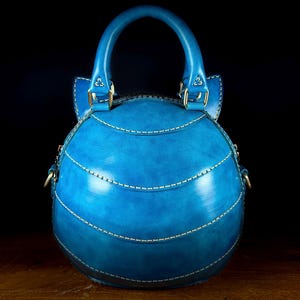 May include: A round, blue leather handbag with a top handle and white stitching. The bag features a curved shape with horizontal stitched lines and gold-colored hardware. The bag is set against a dark background.