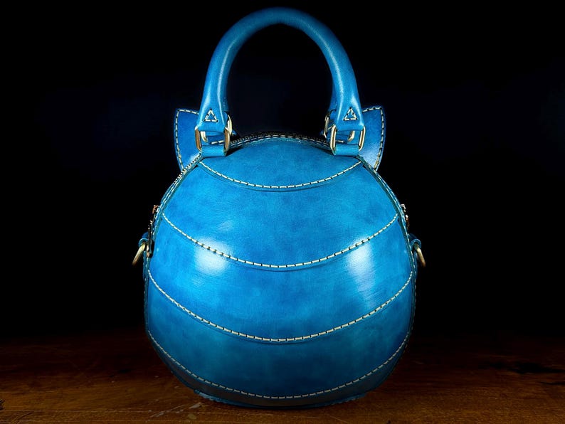 May include: A round, blue leather handbag with a top handle and white stitching. The bag features a curved shape with horizontal stitched lines and gold-colored hardware. The bag is set against a dark background.