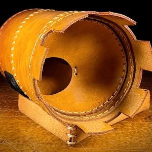 May include: A cylindrical leather object in tan and brown hues, featuring visible stitching and a circular opening. The leather has a textured appearance and is hand-stitched with a lighter thread. It rests on a small, rectangular base.